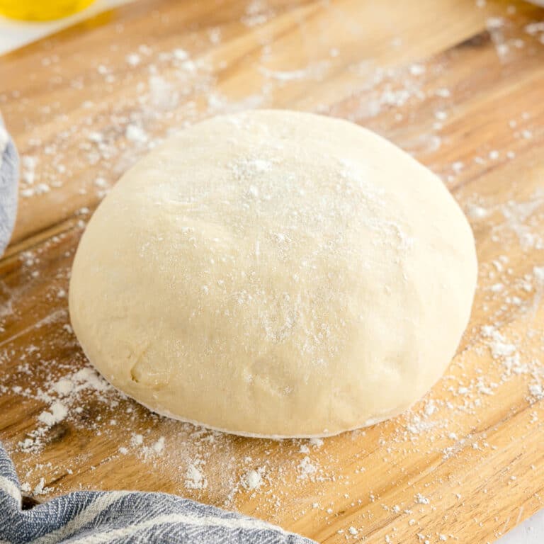 A round ball of raw Pizza dough dusted with flour sits on a floured wooden surface next to a striped cloth.