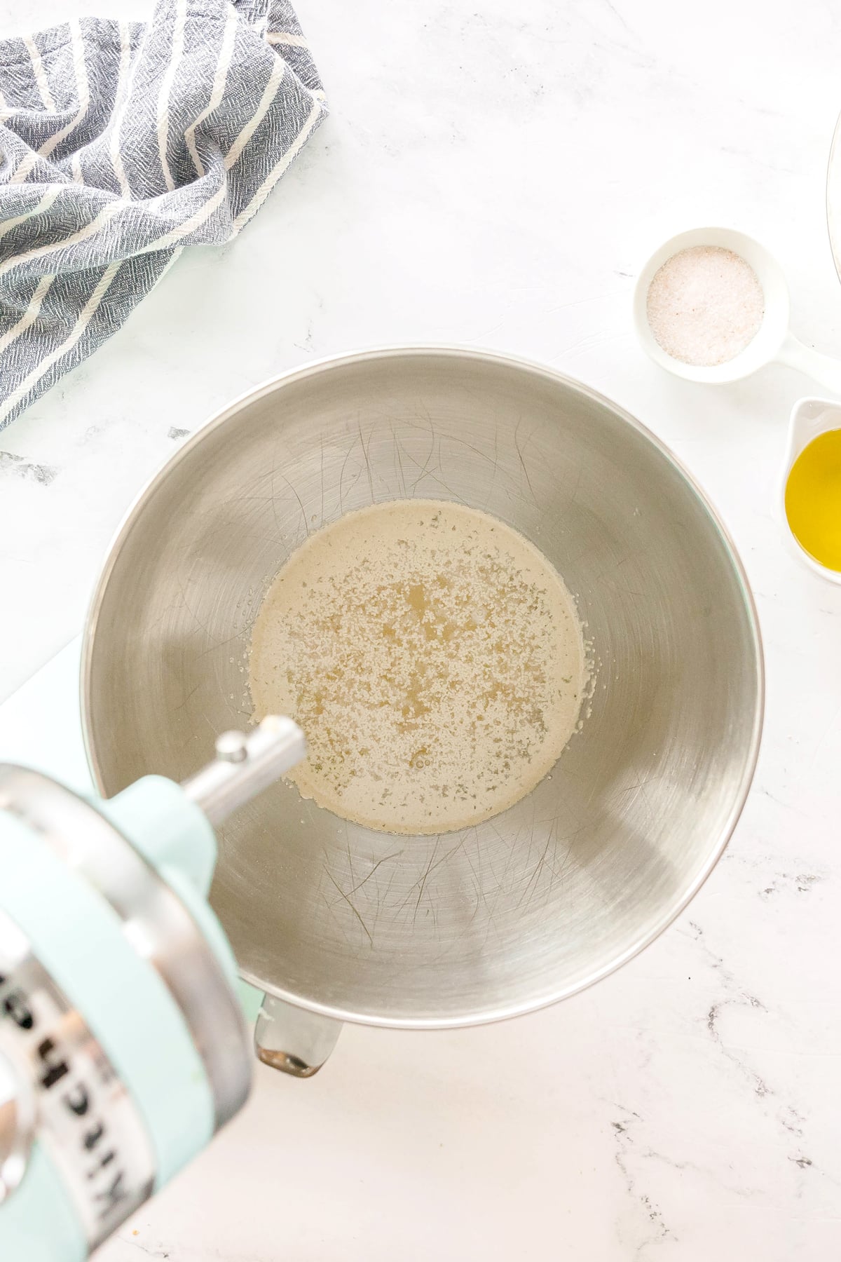 Adding warm water to the bowl of a stand mixer, sprinkle the yeast over it, whisk to combine, and let it rest for about 10 minutes until bubbles form is first step in preparing Pizza Dough
