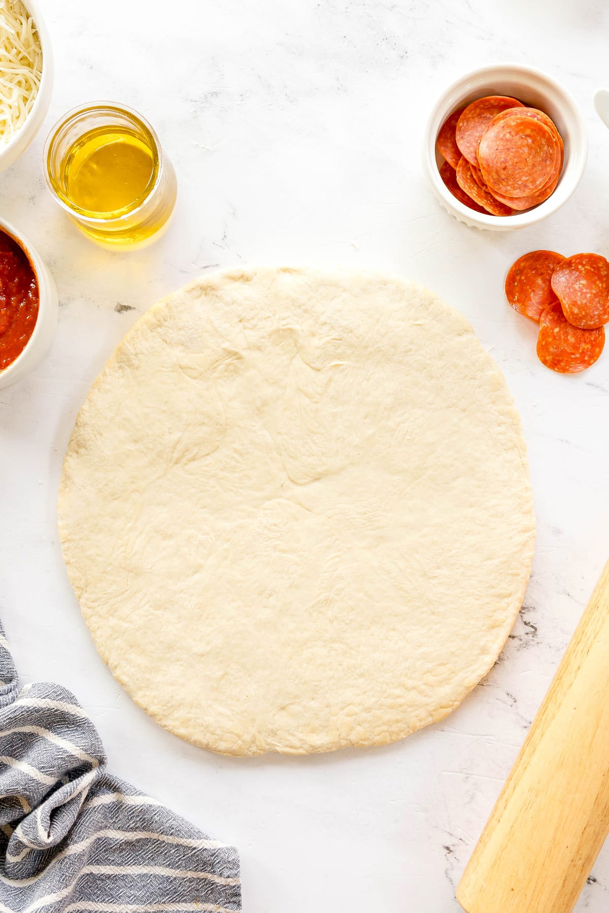 Uncooked pizza dough on a floured surface, surrounded by bowls of olive oil, pizza sauce, shredded cheese, and pepperoni slices.
