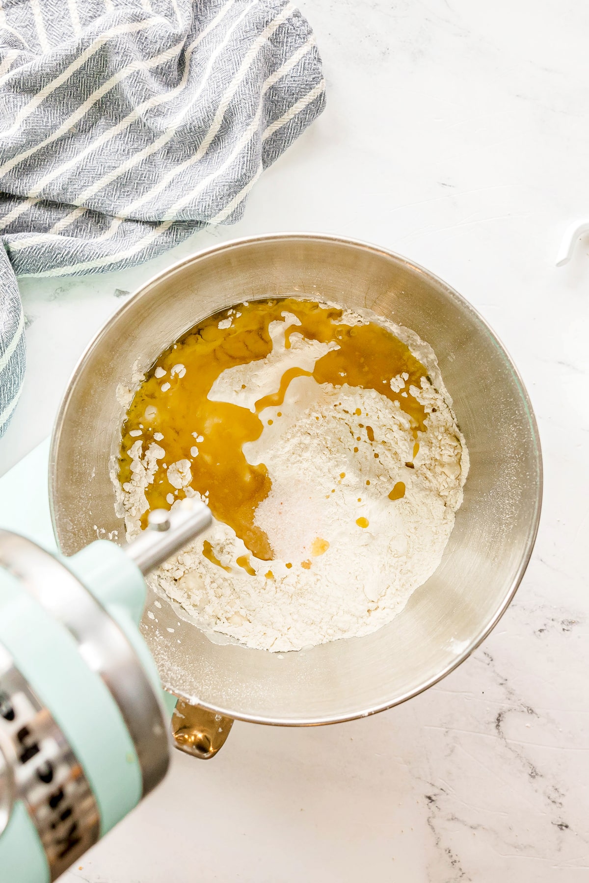 Adding virgin olive oil ito a mixing bowl with flour and liquid ingredients sits under a stand mixer, ready to be mixed. A striped kitchen towel is nearby on a marble countertop. This is one step in preparing Pizza Dough.