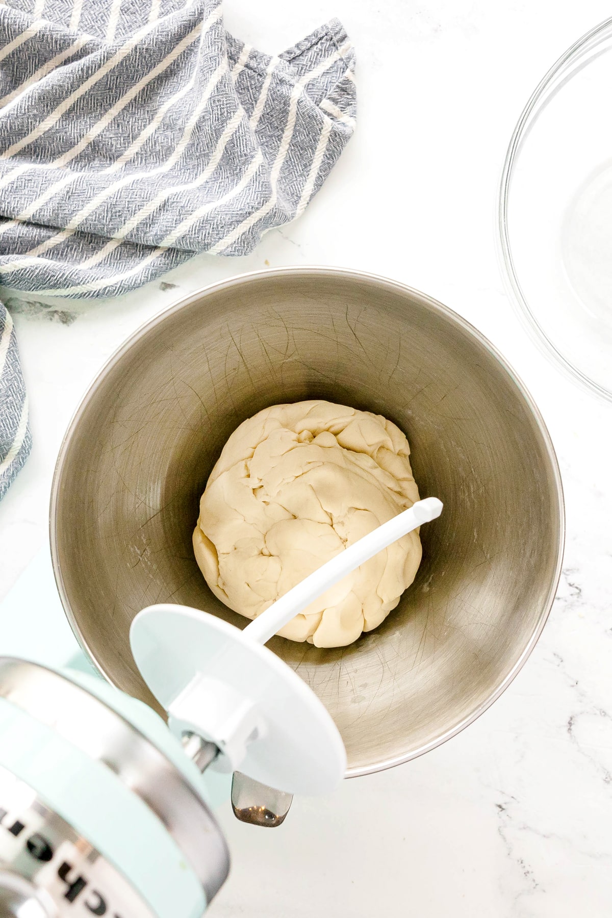 Ball of Pizza dough in a metal mixing bowl with a stand mixer attachment, striped dish towel and glass bowl nearby on a white surface.
