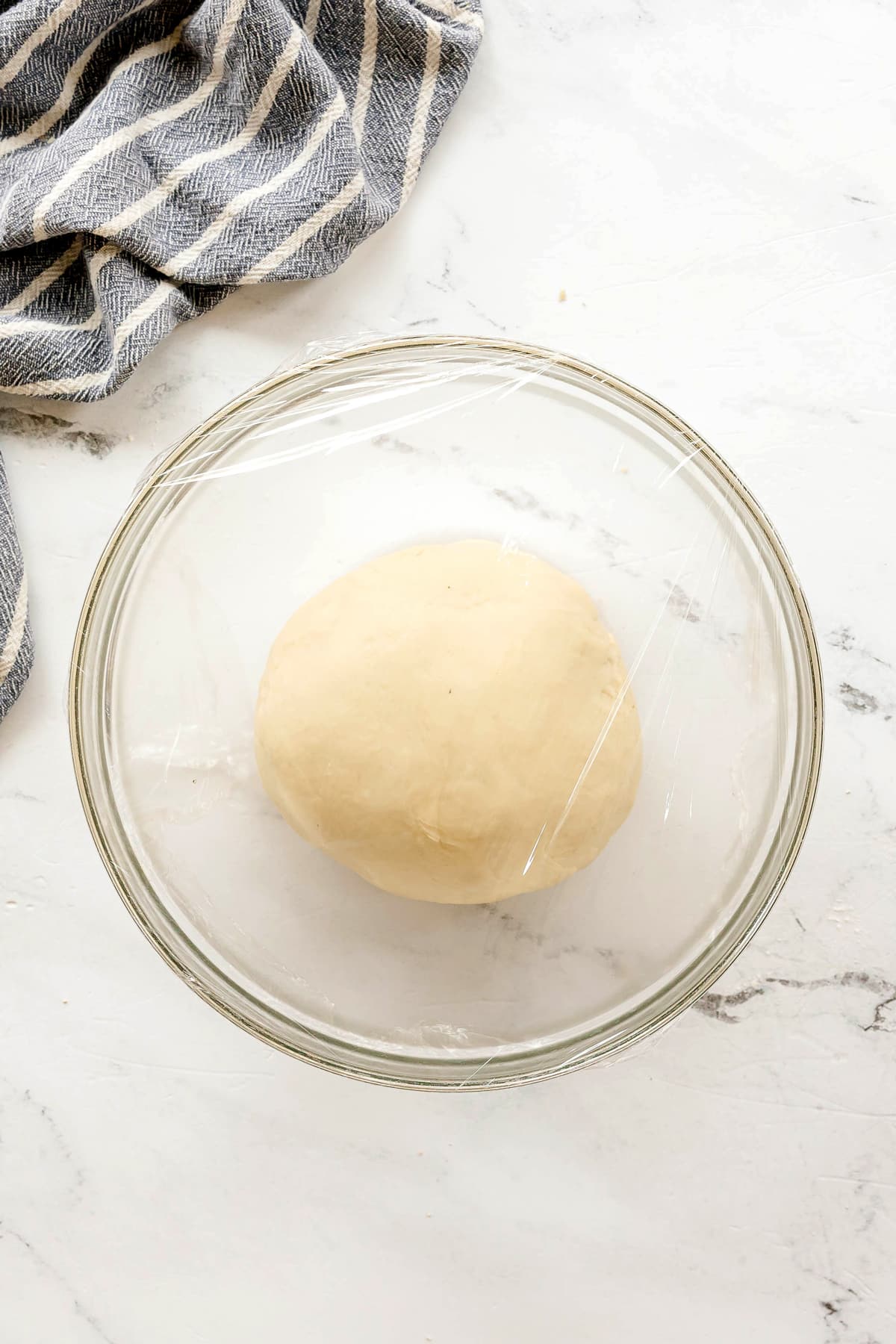 A ball of Pizza Dough sits in a clear glass bowl covered with plastic wrap on a white marble surface next to a striped kitchen towel.