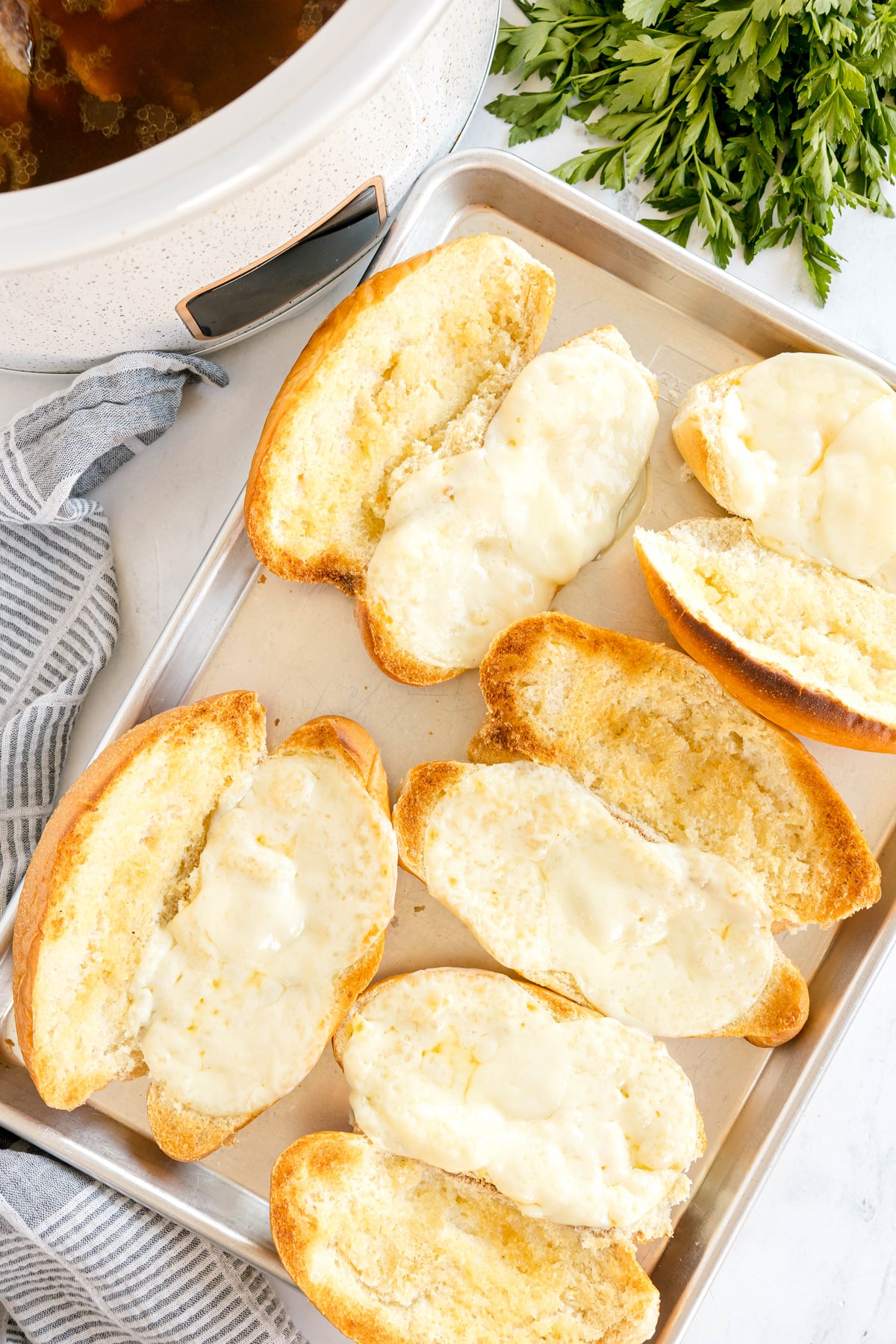 Toasted sandwich rolls with melted cheese on a baking sheet, next to a bunch of fresh parsley and a slow cooker. This is for Slow Cooker French Dip Sandwiches recipe.