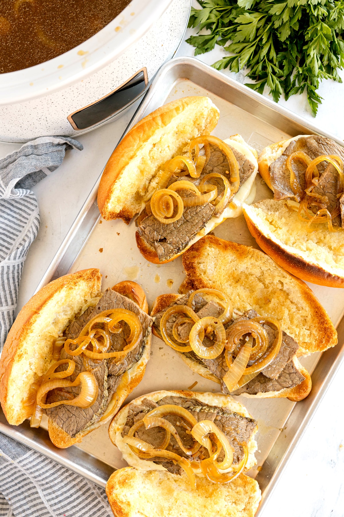 Slow Cooker French Dip Sandwiches with sliced beef and cooked onions on toasted buns, arranged on a baking sheet near a slow cooker and fresh parsley.