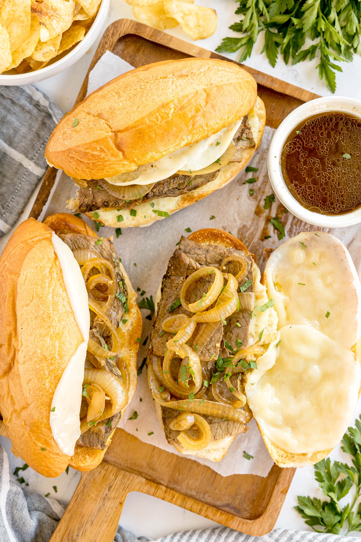 Three French dip sandwiches with melted cheese and sautéed onions on a wooden board, served with a bowl of au jus and garnished with fresh parsley.