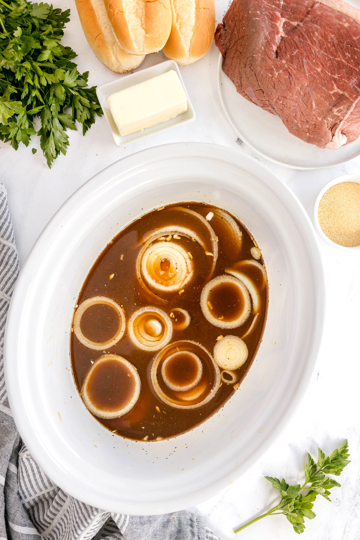 An oval slow cooker filled with broth and sliced onions, and garlic, surrounded by raw beef, buns, butter, parsley, and a small bowl of seasoning on a white surface. This is one of the steps in preparing Slow Cooker French Dip Sandwiches