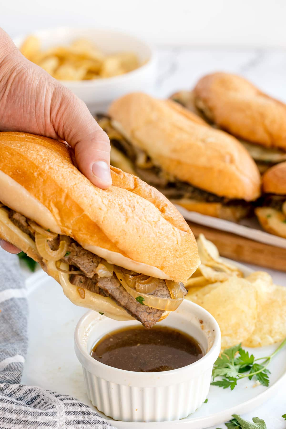 A hand holds Slow Cooker French Dip Sandwich with onions over a ramekin of au jus, with potato chips and more sandwiches in the background.