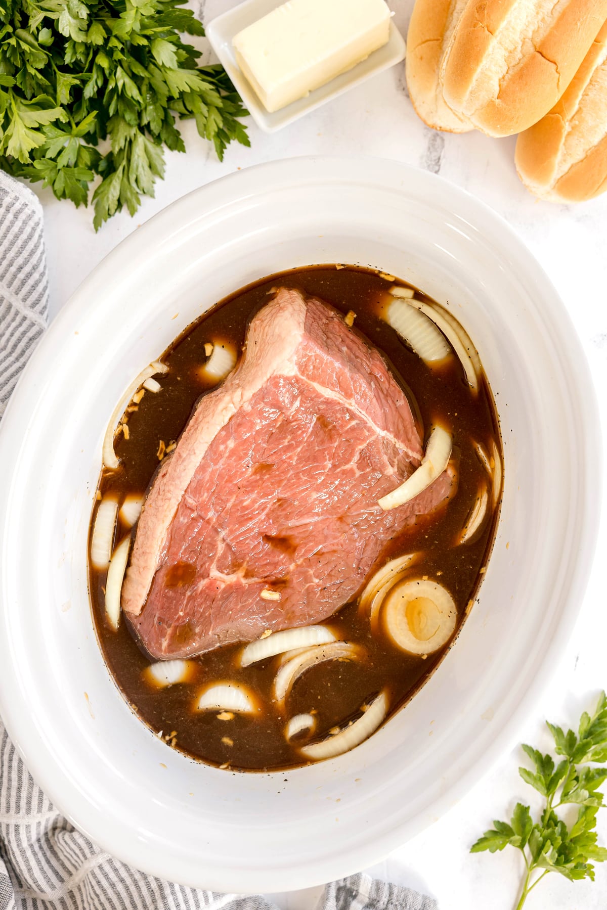 A raw beef roast sits in a white slow cooker with sliced onions and broth for Slow Cooker French Dip Sandwiches. Butter, sandwich rolls, and fresh parsley are nearby.