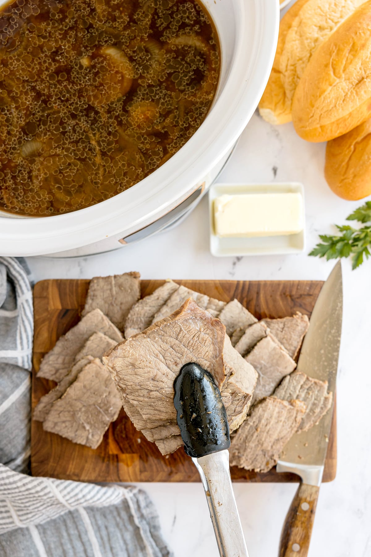 Sliced cooked beef on a wooden board with a knife and tongs, a slow cooker of broth, butter, sandwich rolls, and herbs nearby. These are ingredients of Slow Cooker French Dip Sandwiches