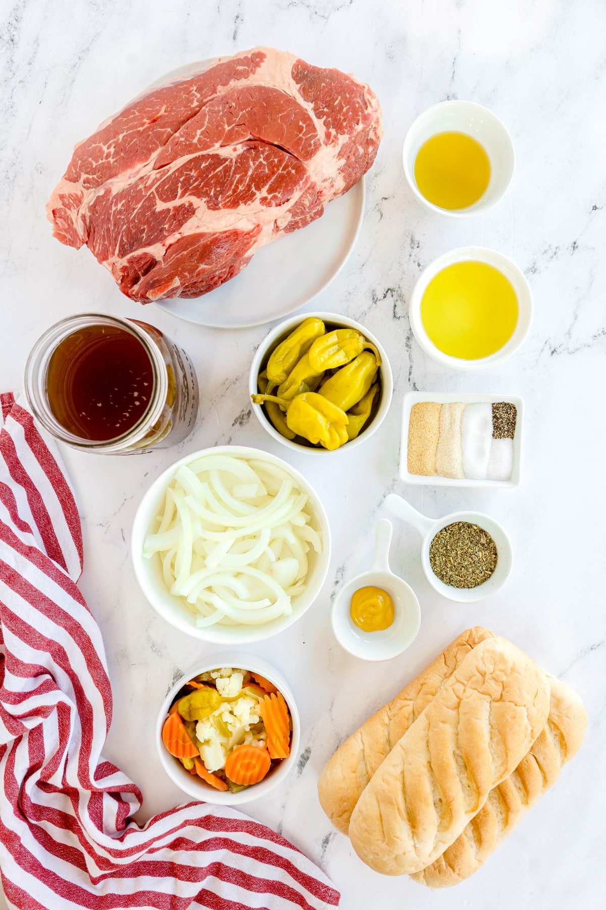 The ingredients for Slow Cooker Italian Beef include beef chuck roast, olive oil, beef broth, pepperoncini juice, whole pepperoncini peppers, onion, Italian seasoning, garlic powder, onion powder, sugar, salt, black pepper, yellow mustard, Italian sandwich rolls, and giardiniera. It is arranged on a white marble counter with stripe linen