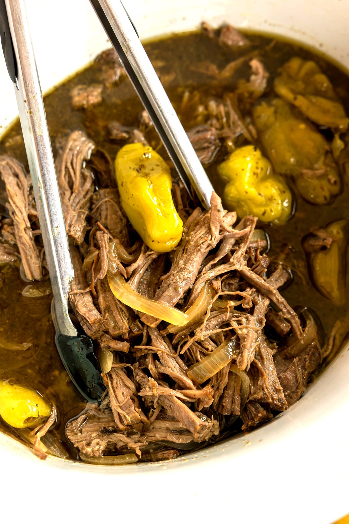 Shredded Slow Cooker Italian Beef with sliced onions and whole pepperoncini peppers in broth, served with metal tongs in a white bowl.