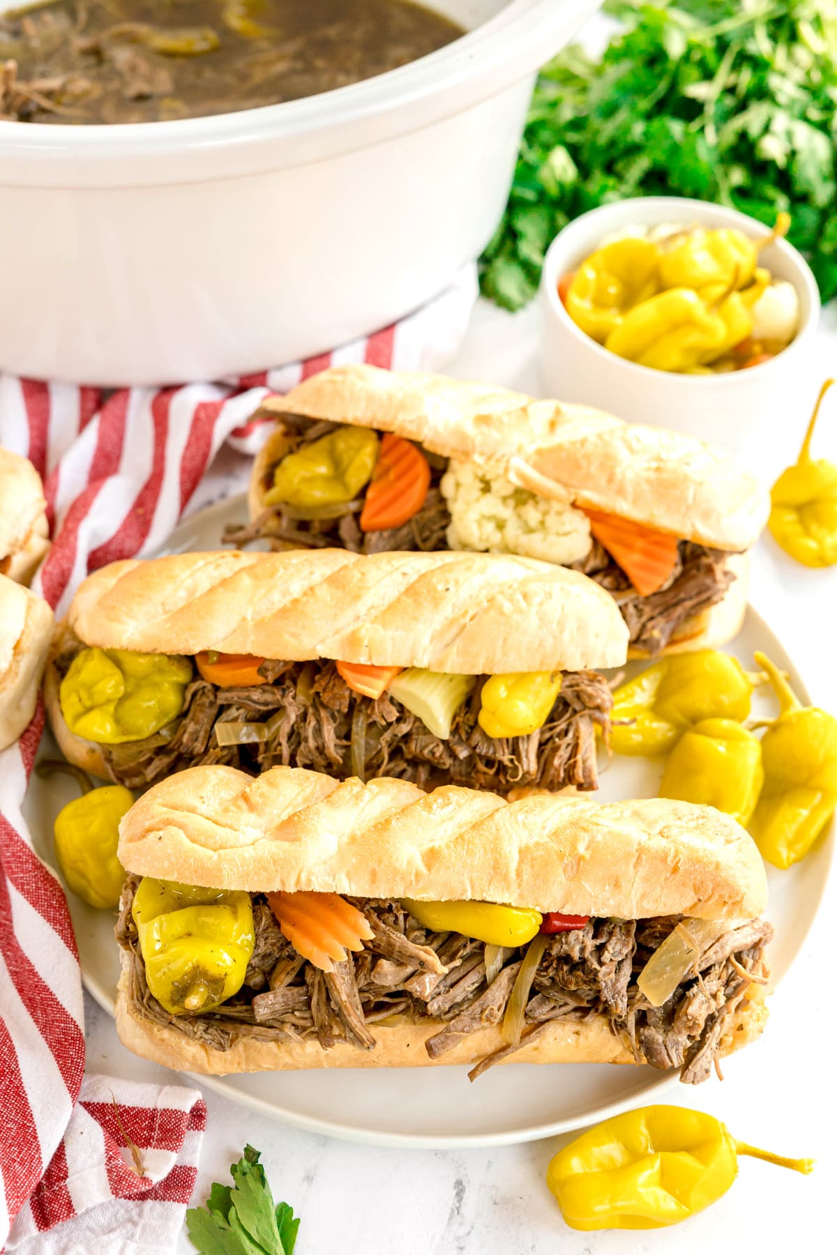 Three Slow Cooker Italian Beef sandwiches with shredded meat, pickled peppers, and vegetables are arranged on a plate, with a small bowl of peppers and a slow cooker in the background.
