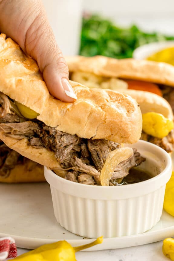 A hand holding a Slow Cooker Italian Beef with onions and peppers, dipping it into a small bowl of au jus, surrounded by more sandwiches and pickled peppers.