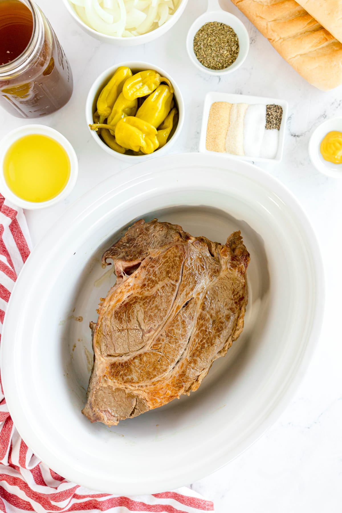 A browned piece of beef sits in a white slow cooker, surrounded by bowls of sliced onions, pepperoncini, dry seasonings, broth, oil, a baguette, and mustard for Slow Cooker Italian Beef recipe