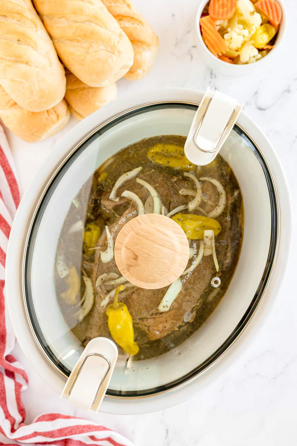 An oval slow cooker with a glass lid contains a roast with sliced onions and peppers and beef. Nearby are three sandwich rolls and a bowl of pickled vegetables. This is one step in preparing Slow Cooker Italian Beef