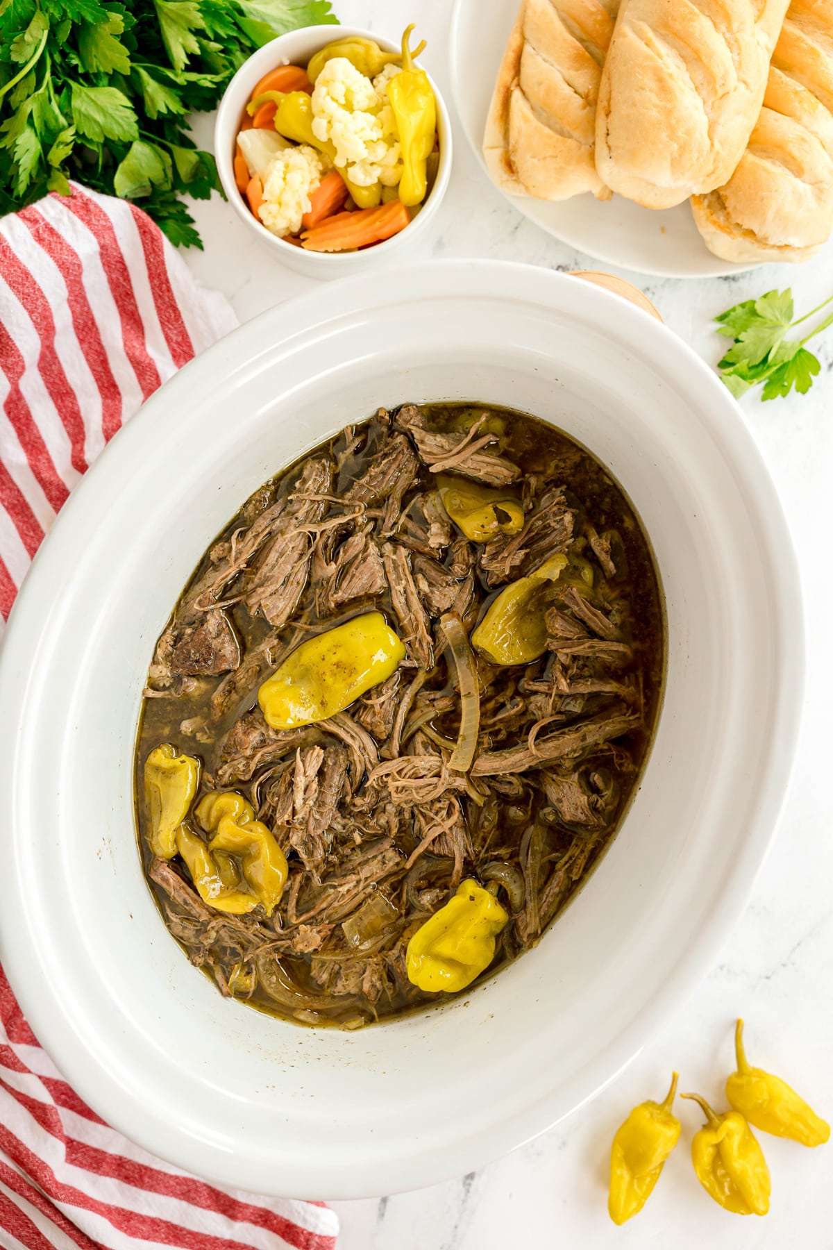 Shredded Slow Cooker Italian Beef with pepperoncini peppers and onions in a white slow cooker, served with sandwich rolls and mixed pickled vegetables on the side.