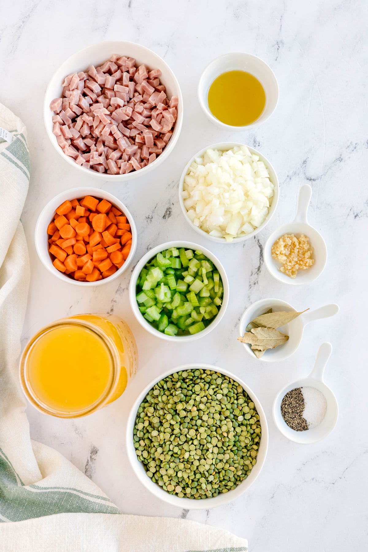 Ingredients for Split Pea and Ham Soup are the following: diced ham, chopped carrots, celery, onions, minced garlic, split peas, chicken broth, olive oil, bay leaves, salt, and pepper are being shown on a marble surface.