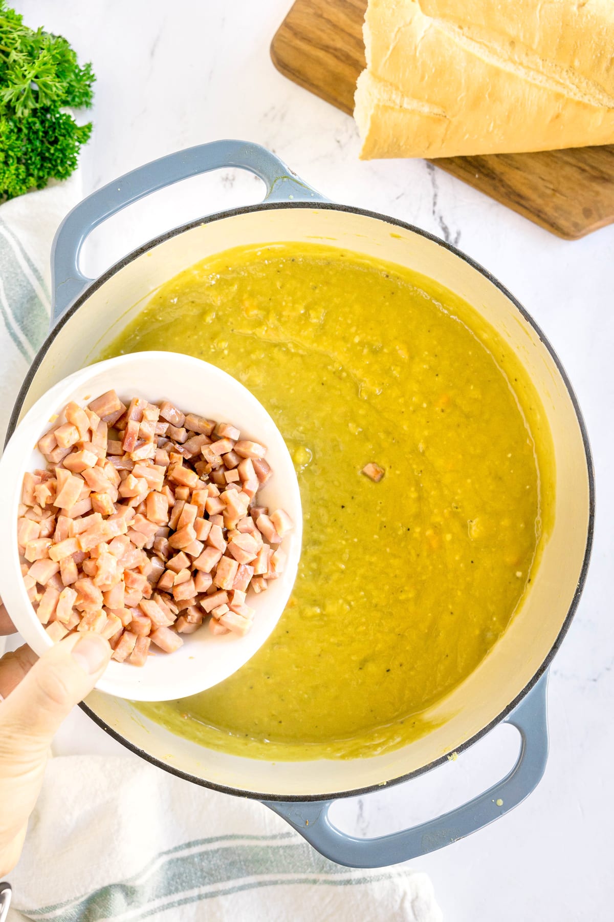 A hand holds a bowl of diced ham over a pot of Split Pea and Ham Soup, ready to add the ham to the soup. A loaf of bread and parsley are in the background.
