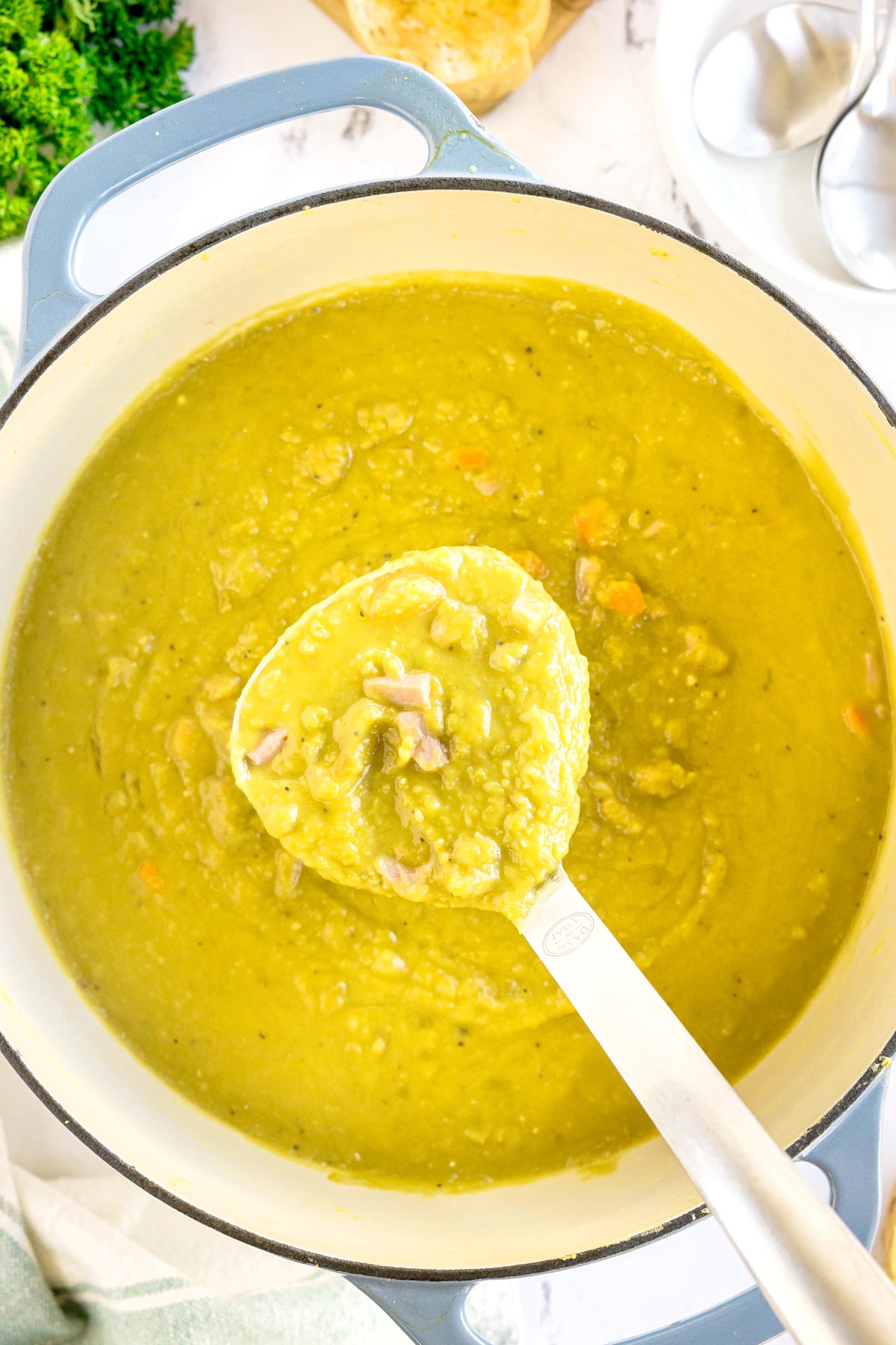 A ladle lifting thick, green split pea soup with chunks of meat and vegetables from a large pot.