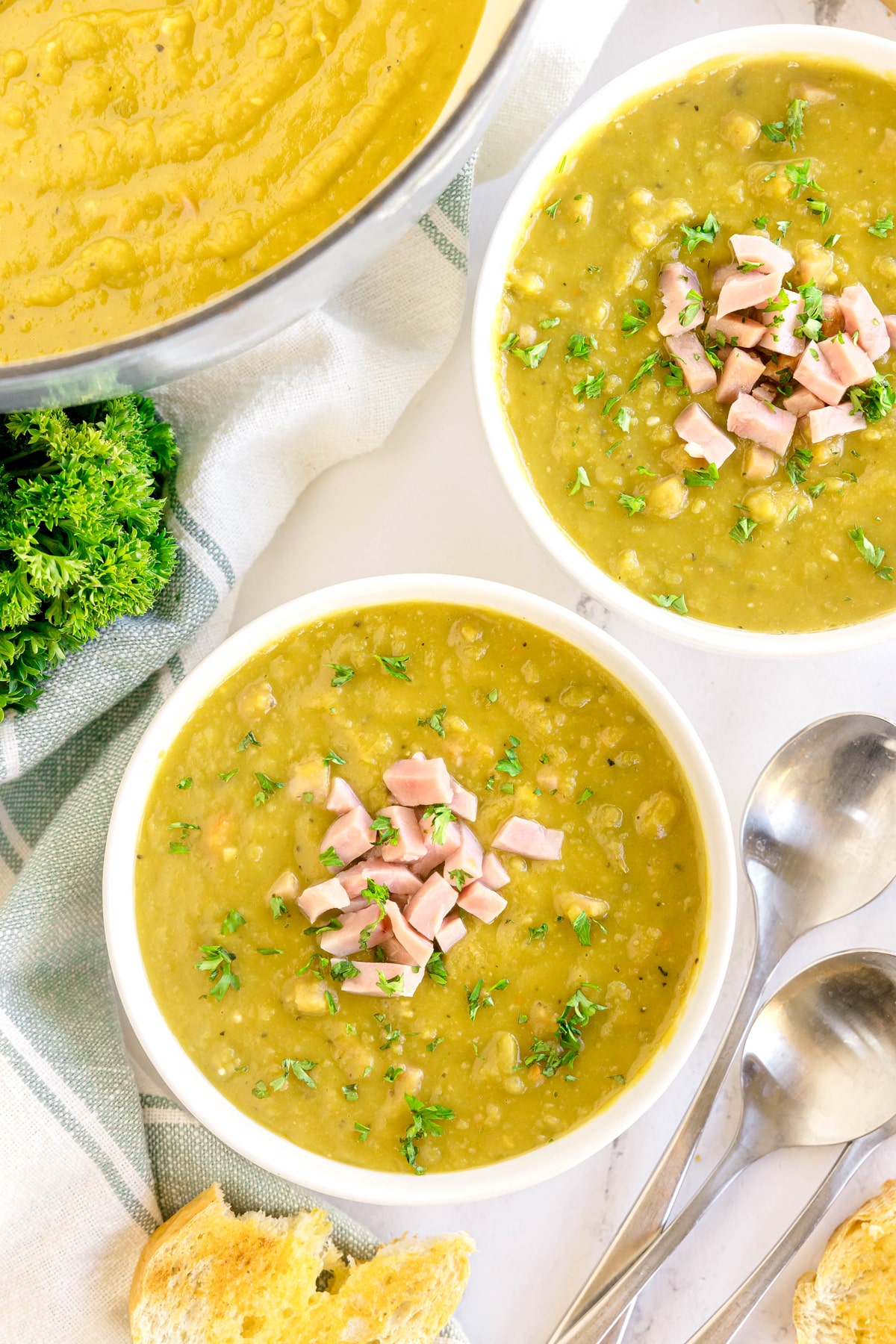 Two bowls of Split Pea and Ham Soup topped with diced ham and chopped parsley, with a pot of soup, fresh parsley, bread, and two spoons nearby.
