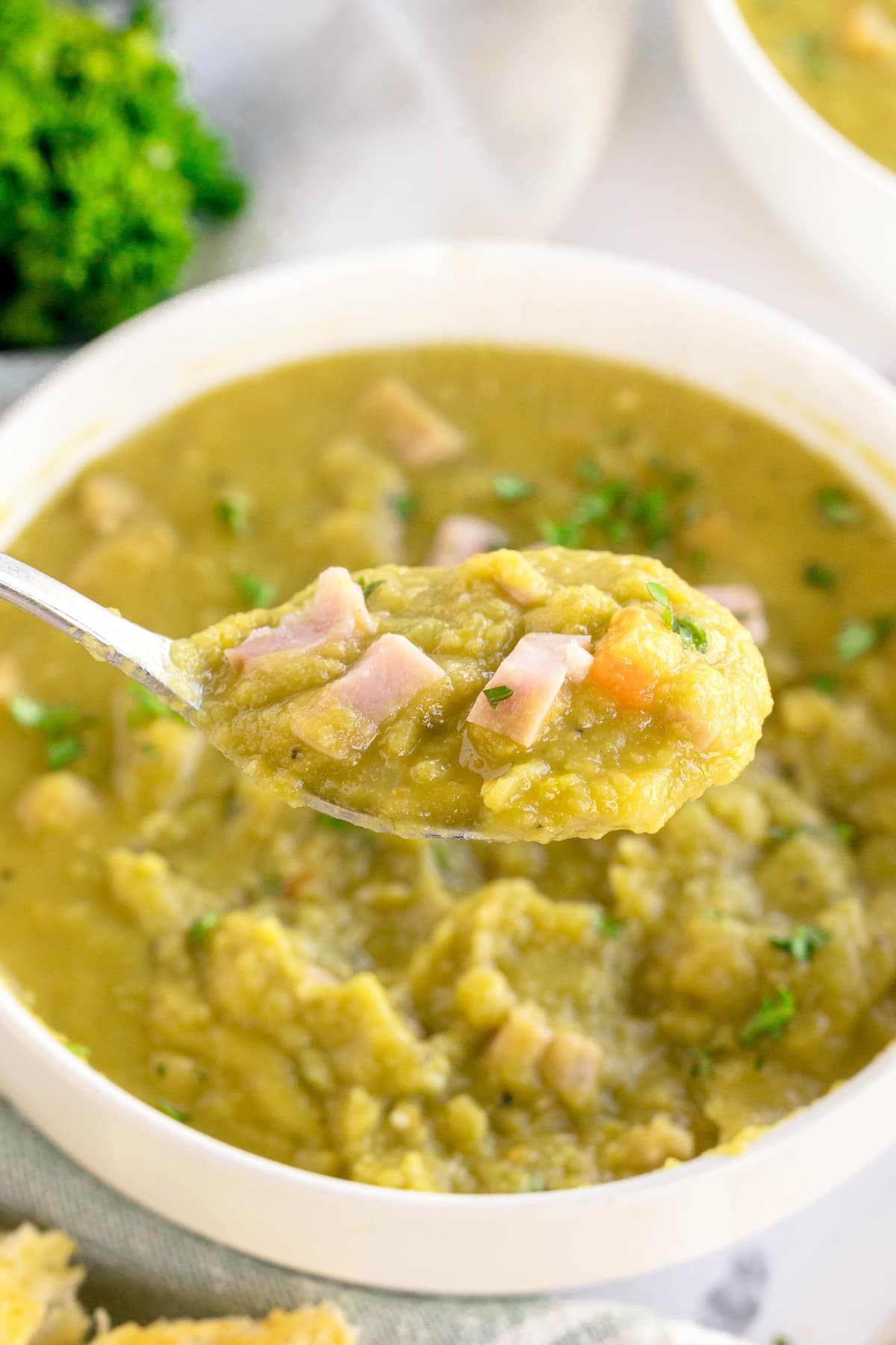 A spoonful of Split Pea and Ham Soup with diced ham is held above a white bowl filled with the same soup.
