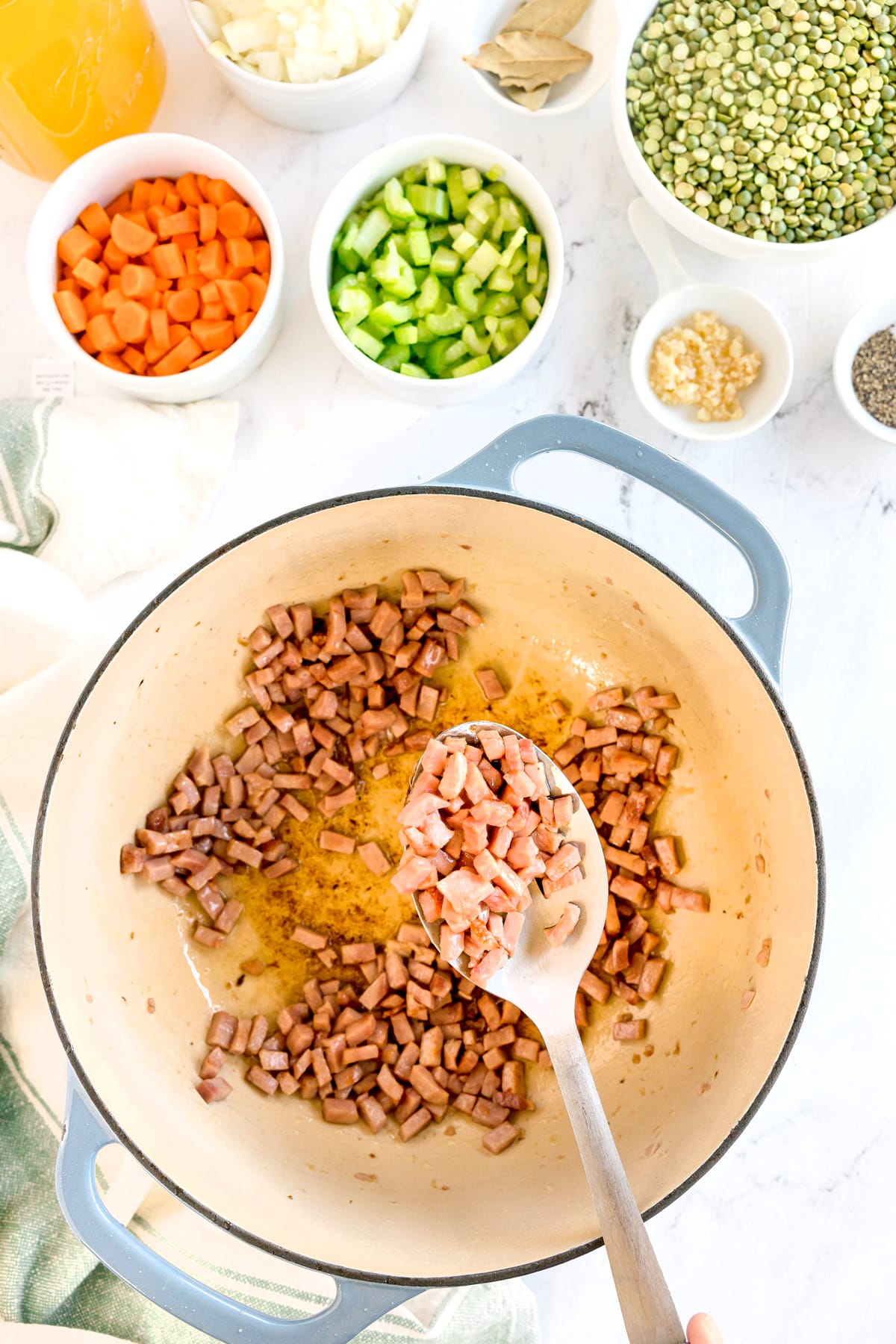 A pot on a stove with diced meat being sautéed, surrounded by bowls of chopped carrots, celery, onions, split peas, garlic, and seasonings. This is first step in preparing Split Pea and Ham Soup
