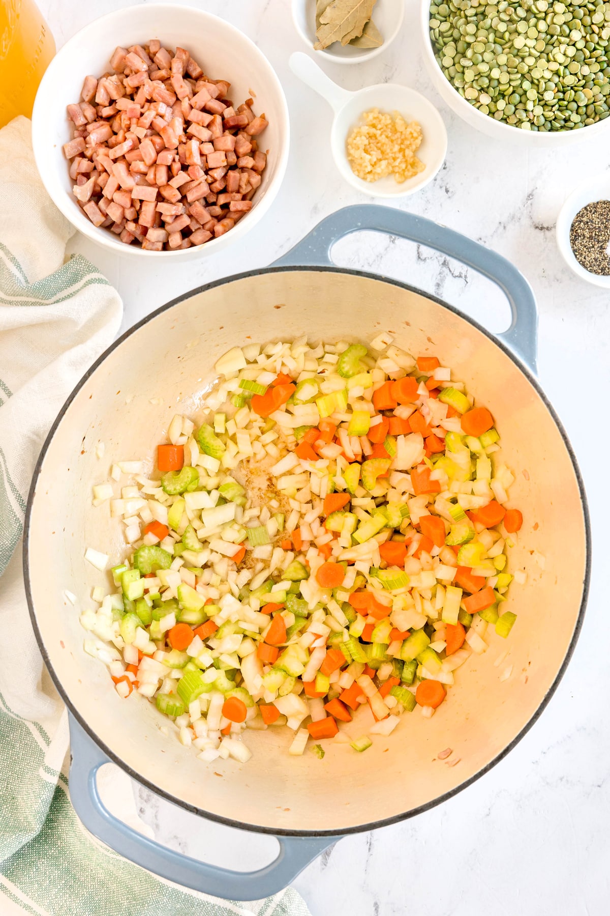 Next step in preparing Split Pea and Ham Soup is to sautee chopped onions, carrots, and celery in a large pot, surrounded by bowls containing diced ham, garlic, seasonings, and split peas on a white countertop.