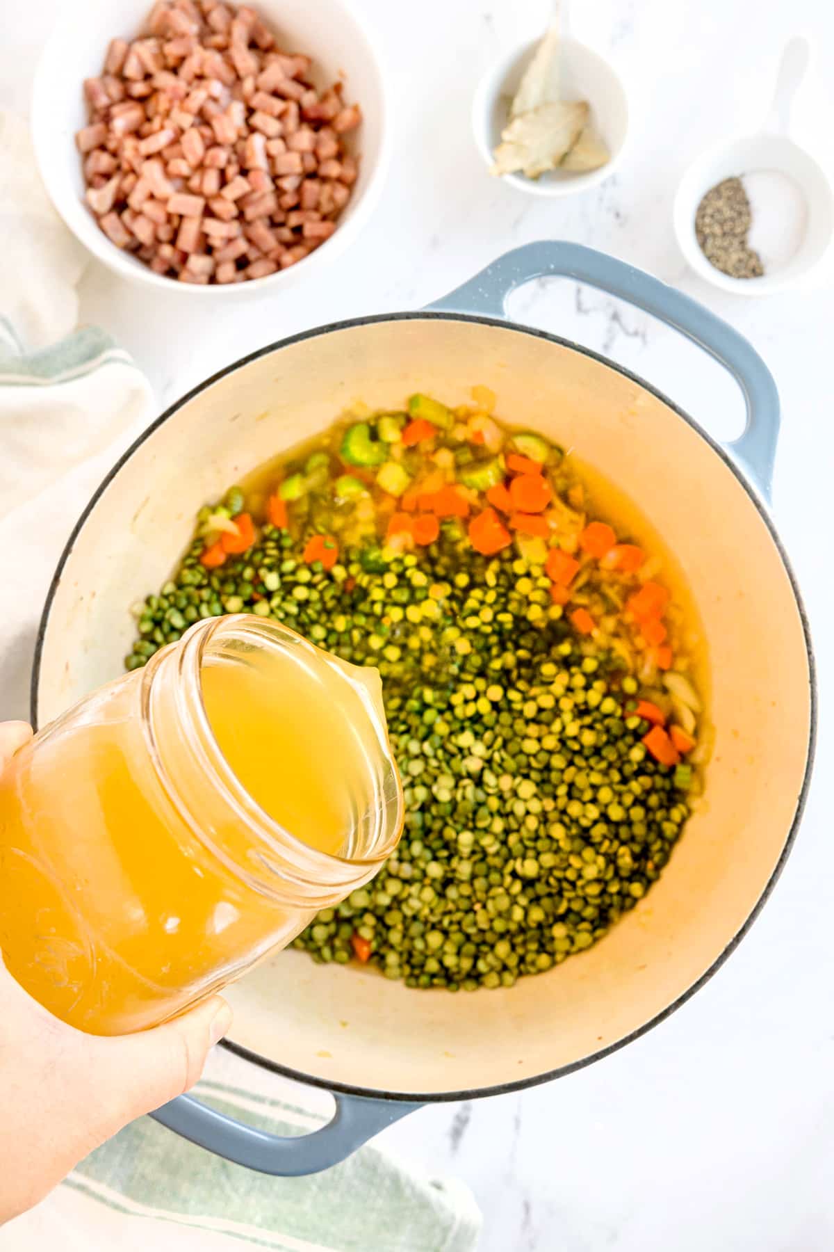 A hand pours broth from a jar into a pot containing split peas, carrots, celery, and onions; diced ham, spices, and a bay leaf are in bowls nearby. This is one of the steps in preparing Split Pea and Ham Soup