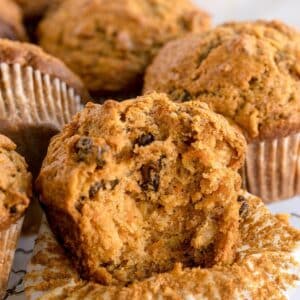 A close-up of several morning glory muffins, with one muffin in front partially broken open to reveal its moist, crumbly texture and visible raisins inside.