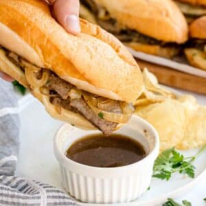 A hand holds a Slow Cooker French Dip Sandwich with sliced beef and onions, dipping it into a ramekin of au jus sauce; potato chips and herbs are on the plate.