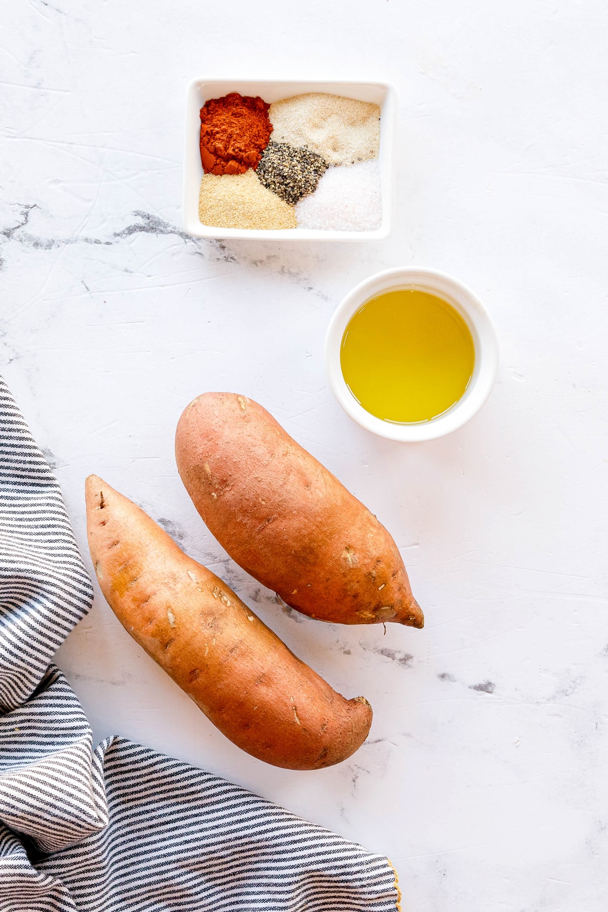 Ingredients for Crispy Air Fryer Sweet Potato Wedges include: two raw sweet potatoes, a small bowl of olive oil, smoked paprika, garlic powder, onion powder, salt, and black pepper, arranged on a white surface next to a striped cloth.