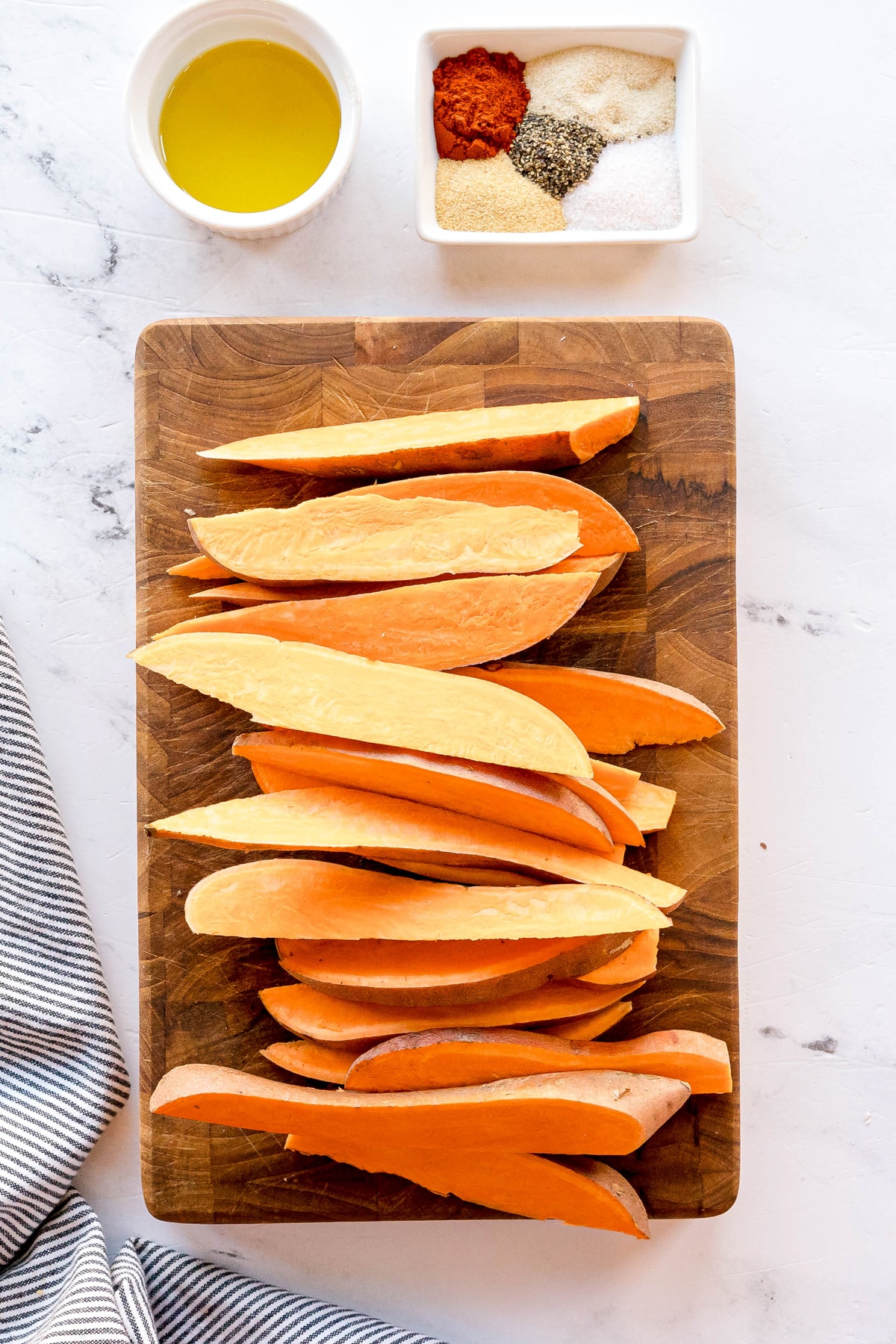 First step in preparing Crispy Air Fryer Sweet Potato Wedges is to slice raw sweet potatoes on a wooden cutting board with a small bowl of oil and a dish of spices on a marble countertop.