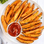 A plate of seasoned potato wedges garnished with herbs, served with a small bowl of ketchup.