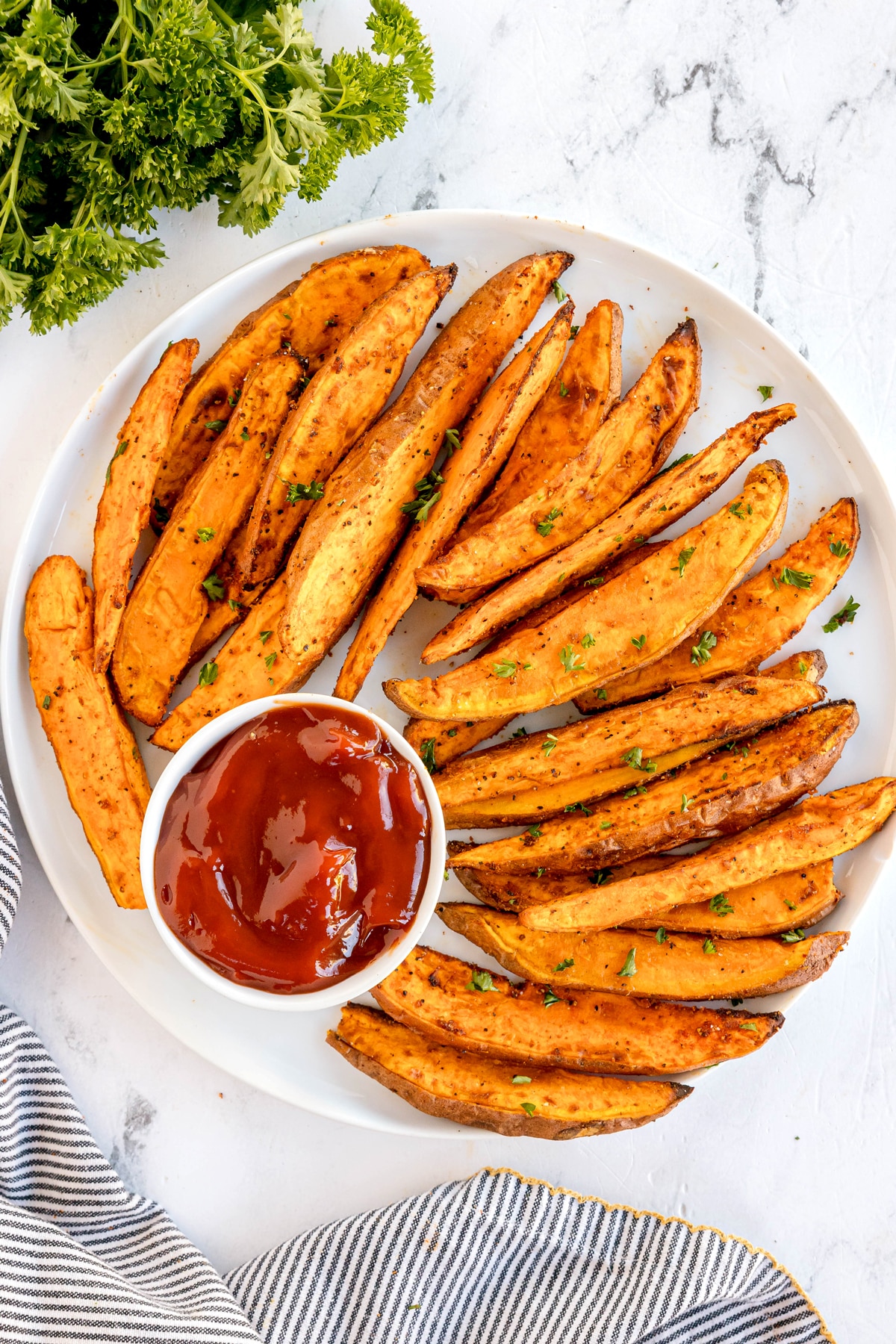 A white plate of seasoned Crispy Air Fryer Sweet Potato Wedges garnished with herbs, served with a small bowl of ketchup on a marble surface.