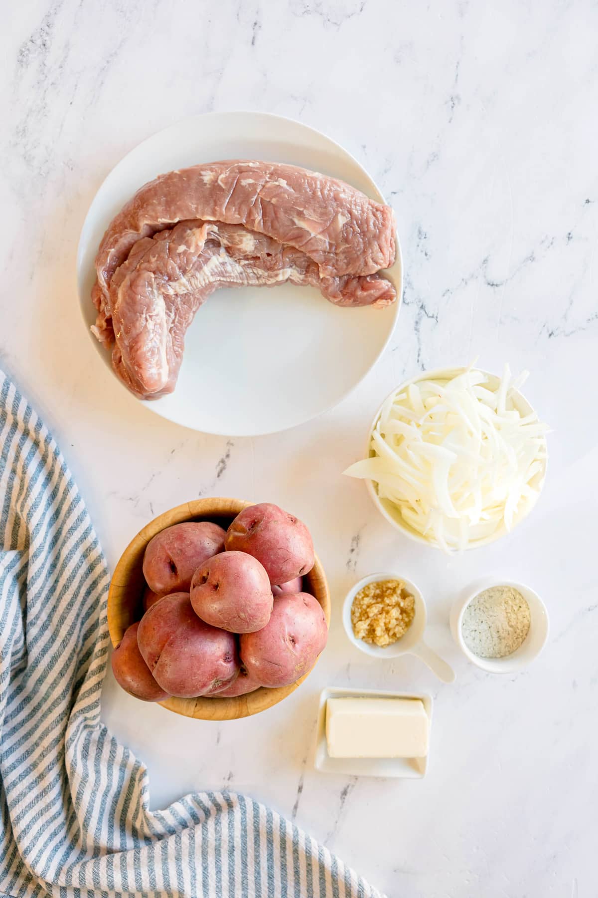 Ingredients on Crockpot Ranch Pork Loin include raw pork tenderloin on a white plate, red potatoes in a wooden bowl, sliced onions, minced garlic, butter, and ranch seasoning on a white marble surface with a striped towel.