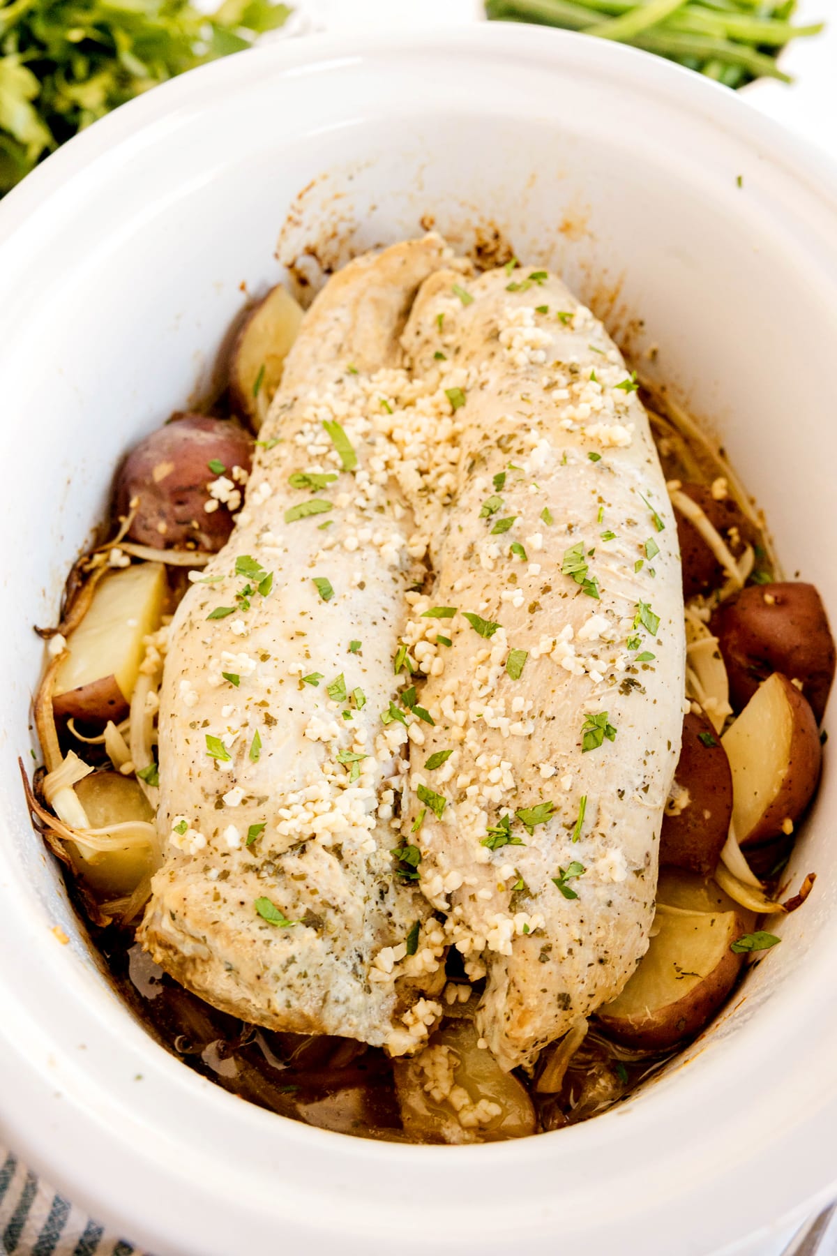 Crockpot Ranch Pork Loin with chopped garlic and herbs sit on top of red potatoes and onions in a white slow cooker.