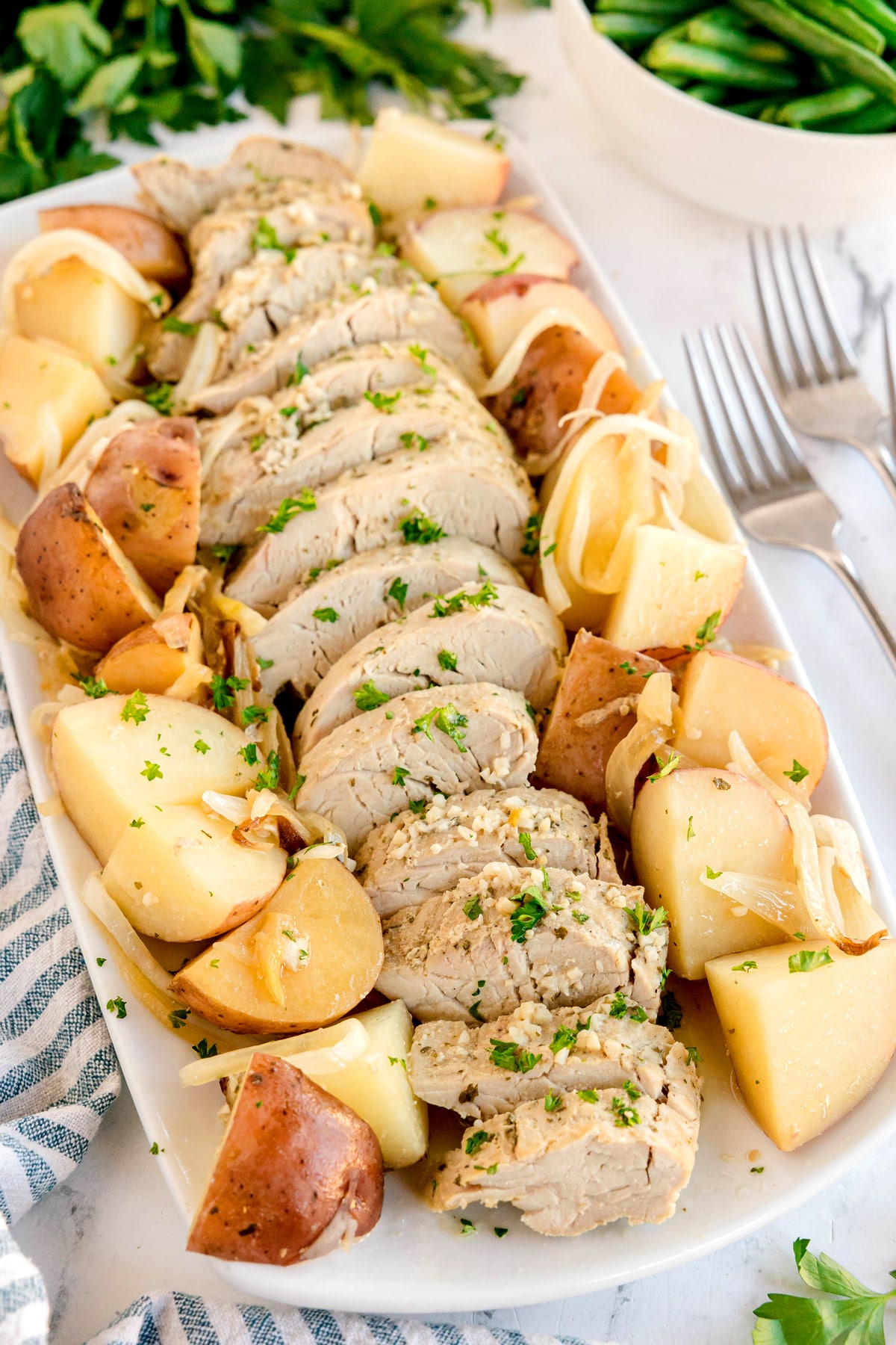 Crockpot Ranch Pork Loin garnished with herbs, surrounded by cooked potato chunks and onions on a rectangular white platter.