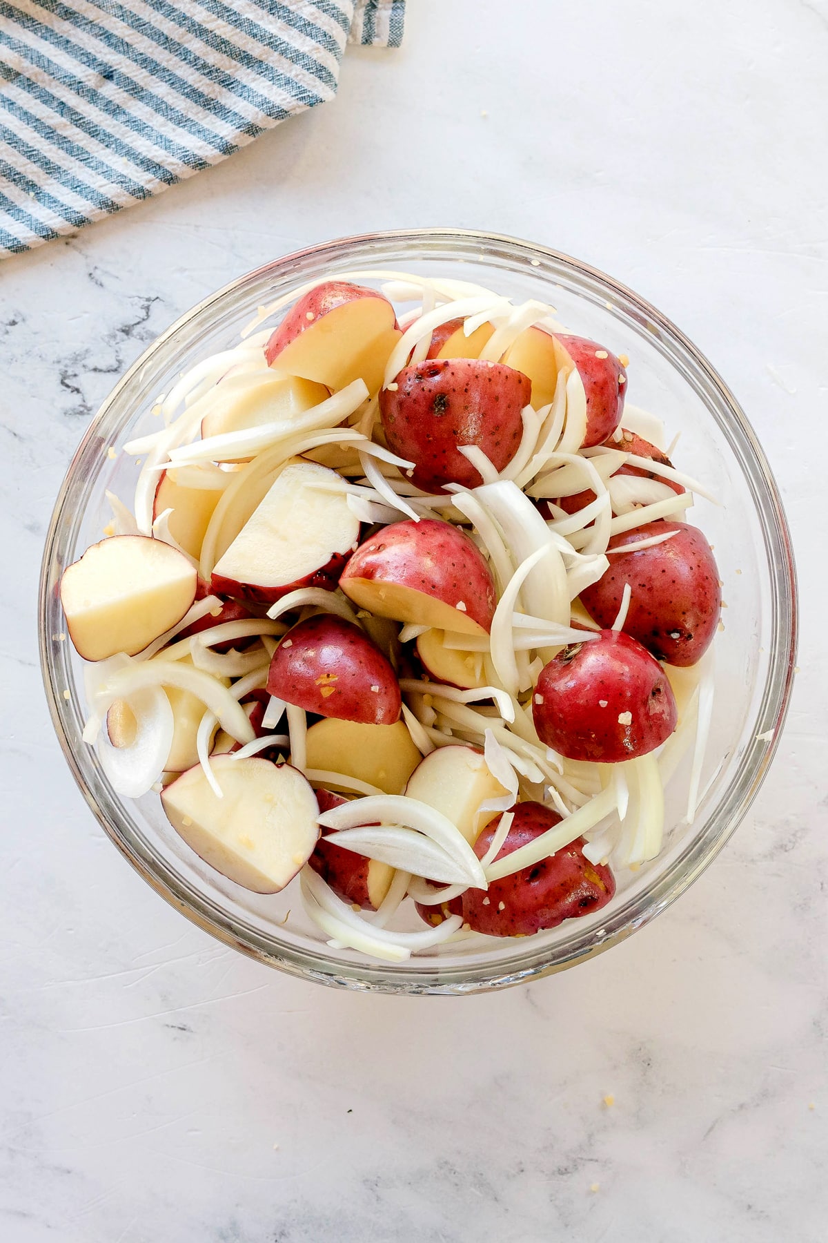 A glass bowl filled with halved red potatoes and sliced onions on a light-colored surface with a striped cloth in the background for Crockpot Ranch Pork Loin recipe