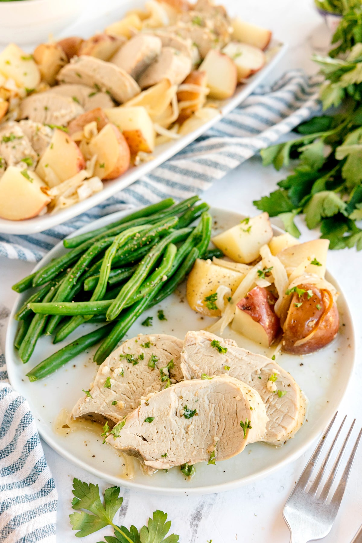 A plate with Crockpot Ranch Pork Loin including green beans, and cooked potatoes. More pork, potatoes, and onions are on a platter in the background with fresh parsley nearby.