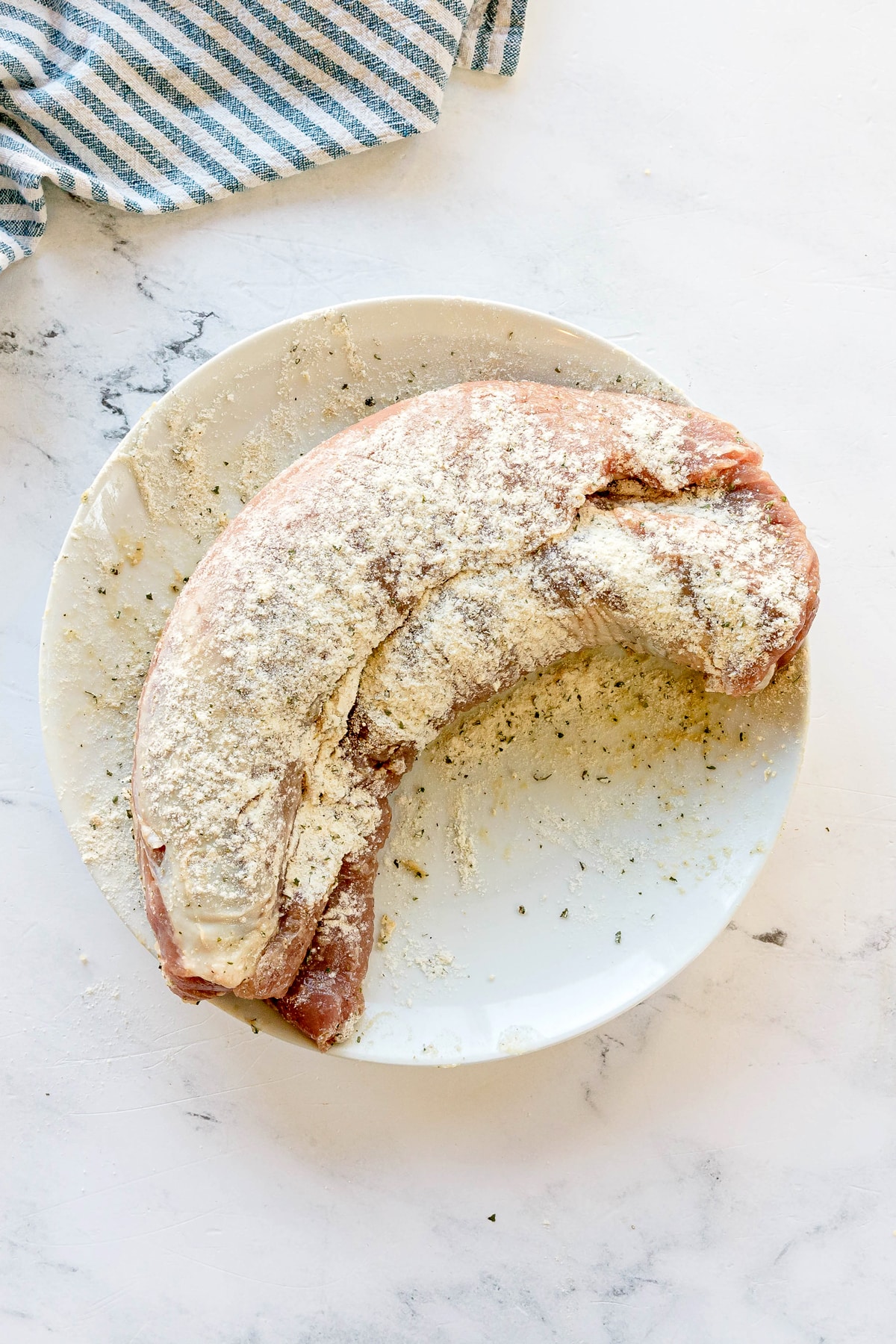 Next step in preparing Crockpot Ranch Pork Loin is to coat raw pork tenderloin coated in 2 tablespoons of ranch seasoning. It sits on a white plate, with a blue and white striped cloth in the background.