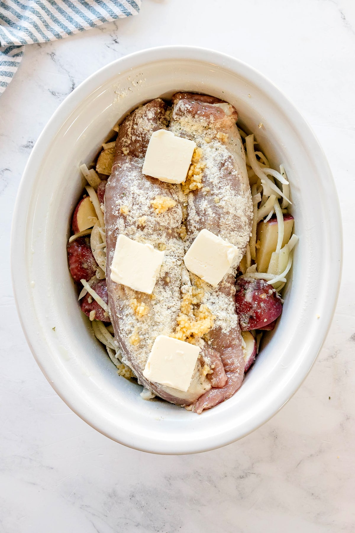 Raw pork tenderloin topped with butter, minced garlic, and seasoning sits atop sliced potatoes and onions in a white slow cooker for Crockpot Ranch Pork Loin recipe.