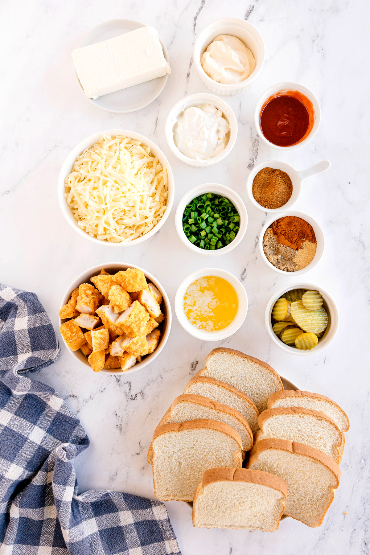 Ingredients for Nashville Hot Chicken Dip recipe arranged on a marble surface, including white bread slices, mozarella cheese, cream cheese, mayonnaise, sour cream, green onions, hot sauce, cayenne pepper, brown sugar, paprika, garlic powder, kosher salt, breaded chicken, and pickle chips. A blue checkered cloth can also be seen on the side.