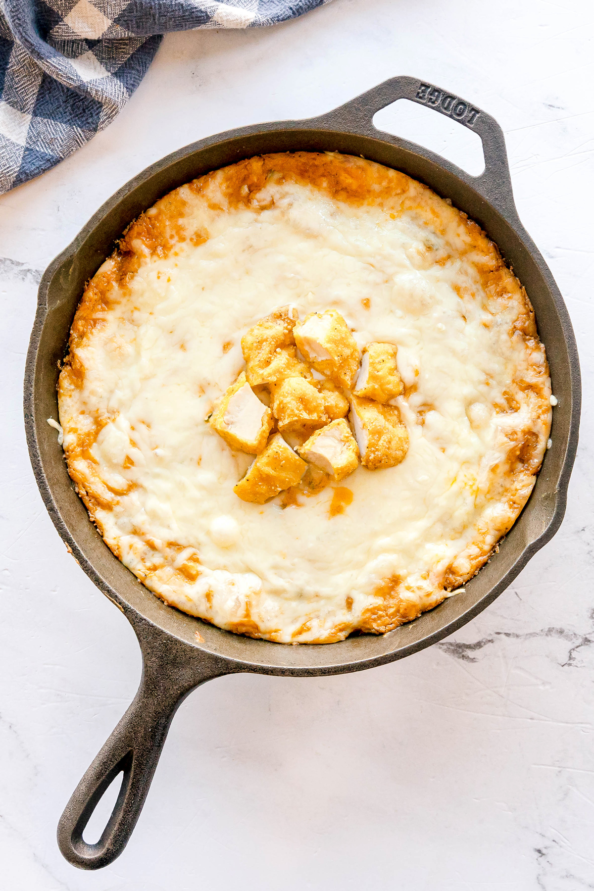 A cast iron skillet filled with Nashville Hot Chicken Dip mixture, topped with melted cheese and chunks of chicken, placed on a marble surface next to a checkered cloth.