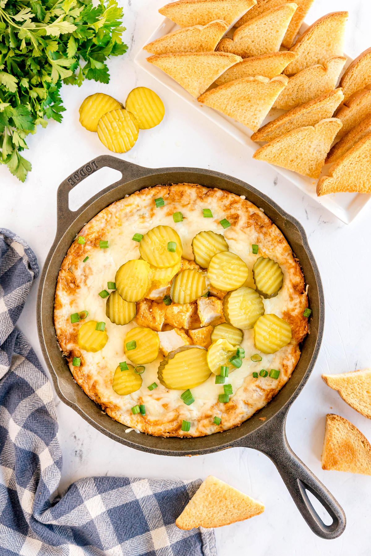 A cast iron skillet containing Nashville Hot Chicken Dip topped with sliced pickles and chopped green onions, surrounded by toasted bread triangles and fresh parsley.