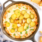 A cast iron skillet filled with a baked Nashville Hot Chicken Dip, topped with sliced pickles and chopped green onions, with bread pieces on the side.