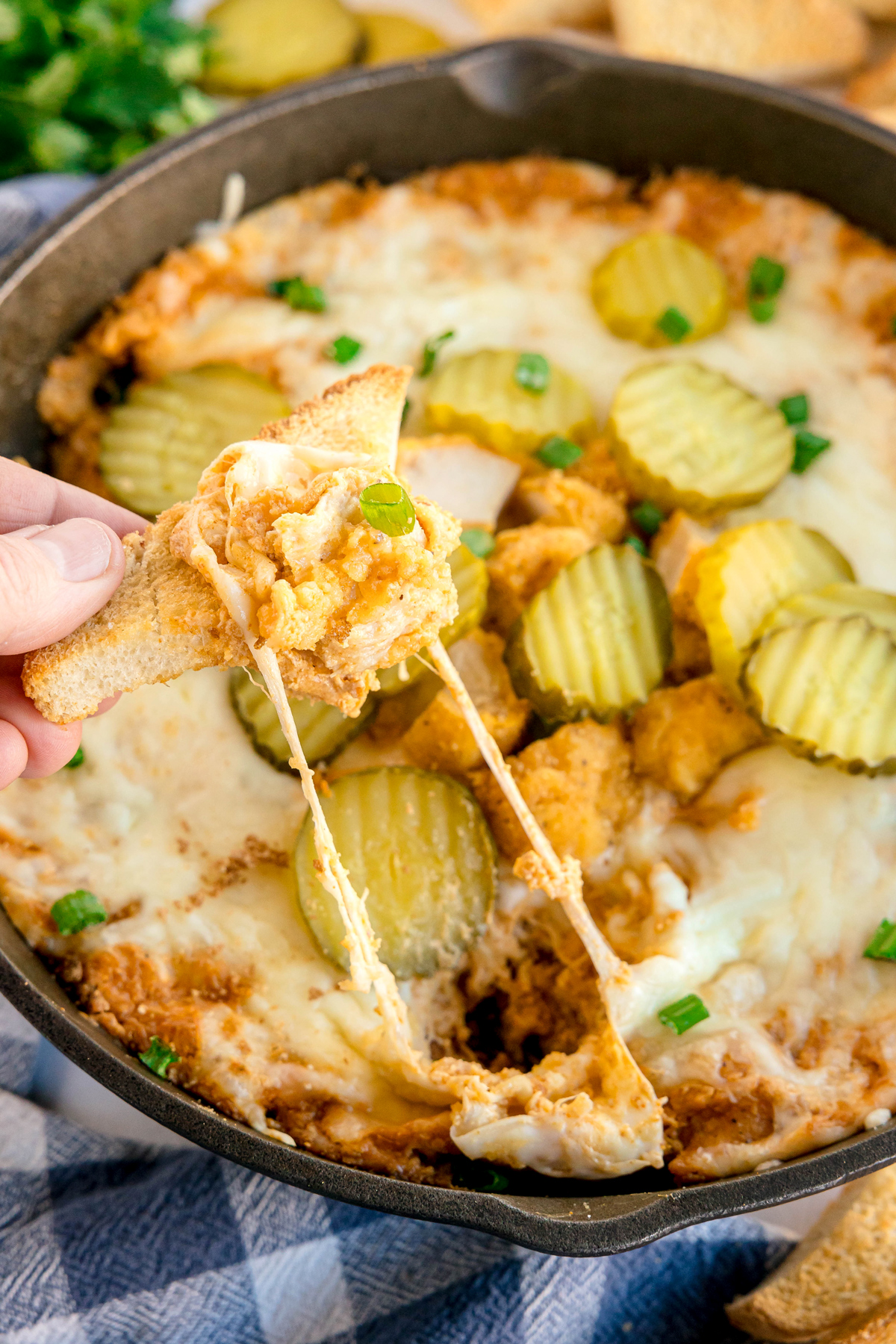 A hand dips a tortilla chip into a skillet filled with a cheesy Nashville Hot Chicken Dip, baked dip topped with sliced pickles and green onions.