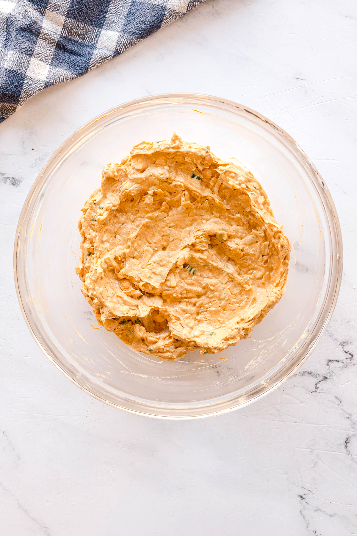 A glass bowl filled with a creamy Nashville Hot Chicken Dip, on a light marble countertop next to a blue and white checkered cloth.