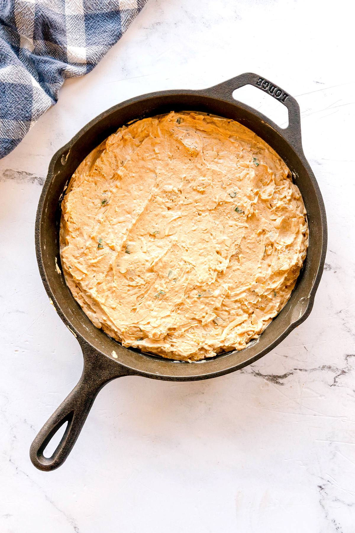 A cast iron skillet filled with Nashville Hot Chicken Dip mixture, placed on a white marble surface next to a blue and white checkered cloth.