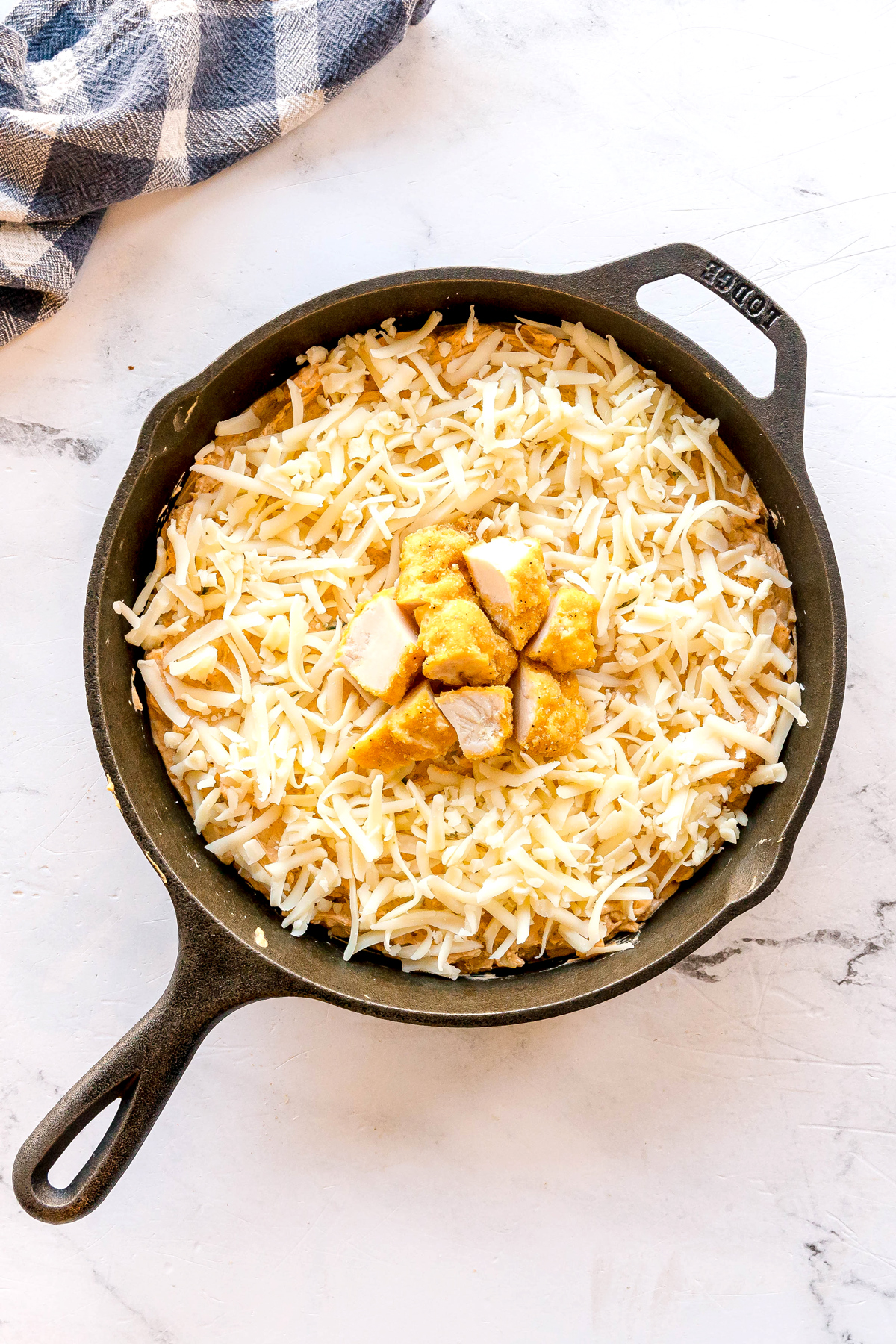 A cast iron skillet filled with shredded cheese and chicken pieces on top. This is one step in preparing Nashville Hot Chicken Dip. Light-colored surface with a checkered cloth nearby can also be seen.