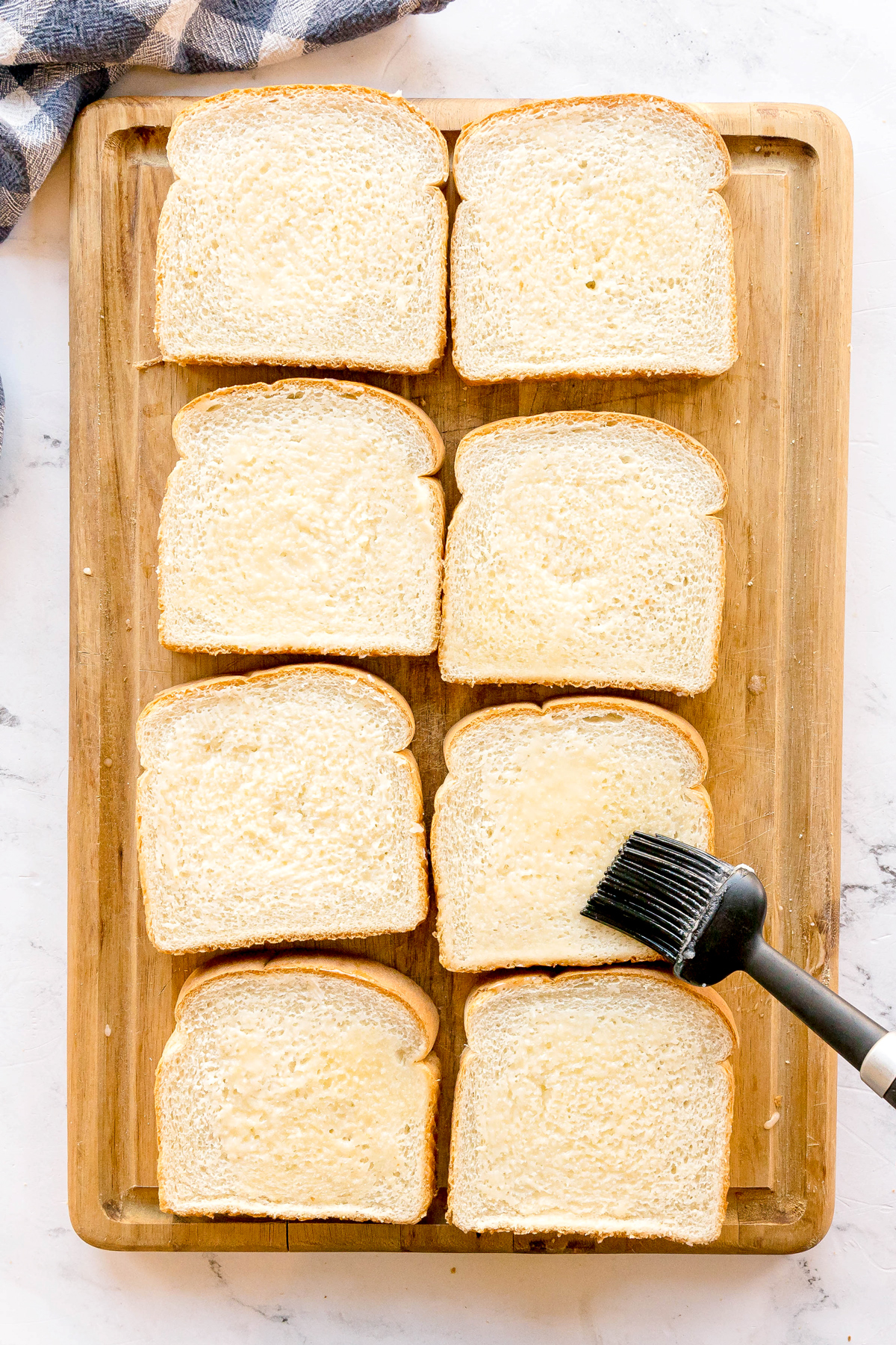 Eight slices of white bread are arranged on a wooden cutting board, with a basting brush applying butter to one slice for Nashville Hot Chicken Dip recipe.